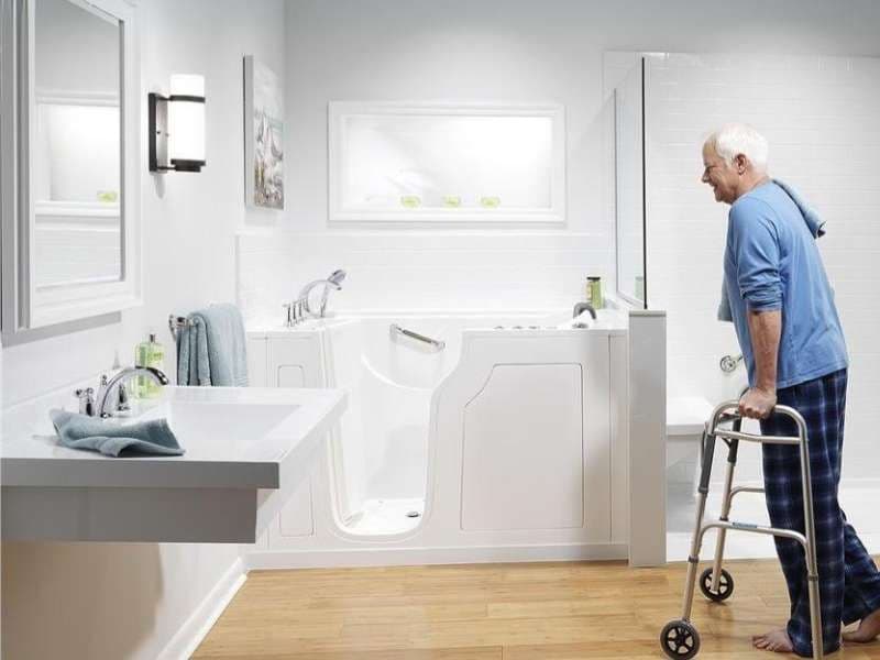 A person uses a walker and approaches an accessible walk-in bathtub in a white bathroom.