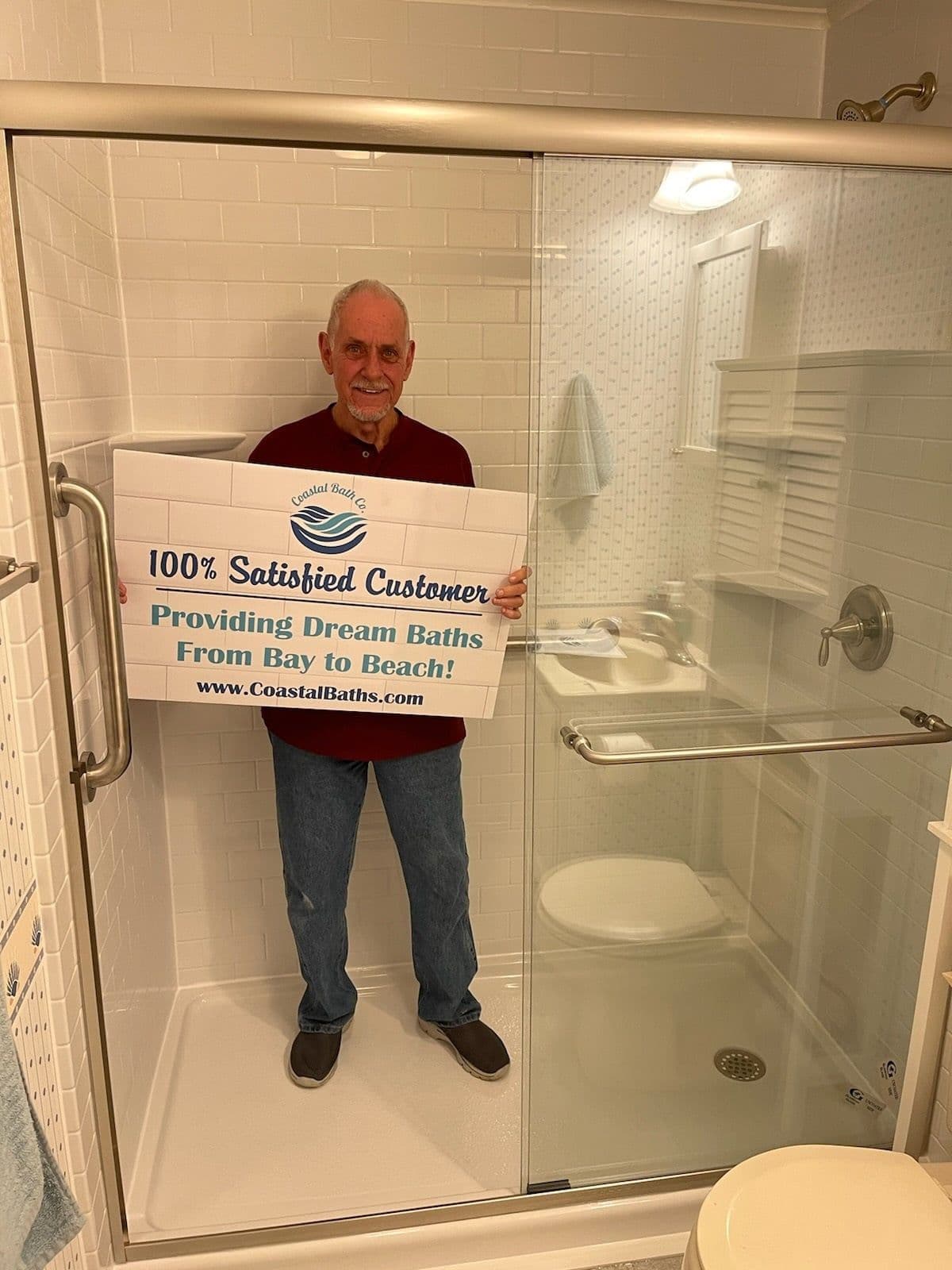 A customer stands in a new walk-in shower with sliding glass doors and holds a sign that reads "100% Satisfied Customer".