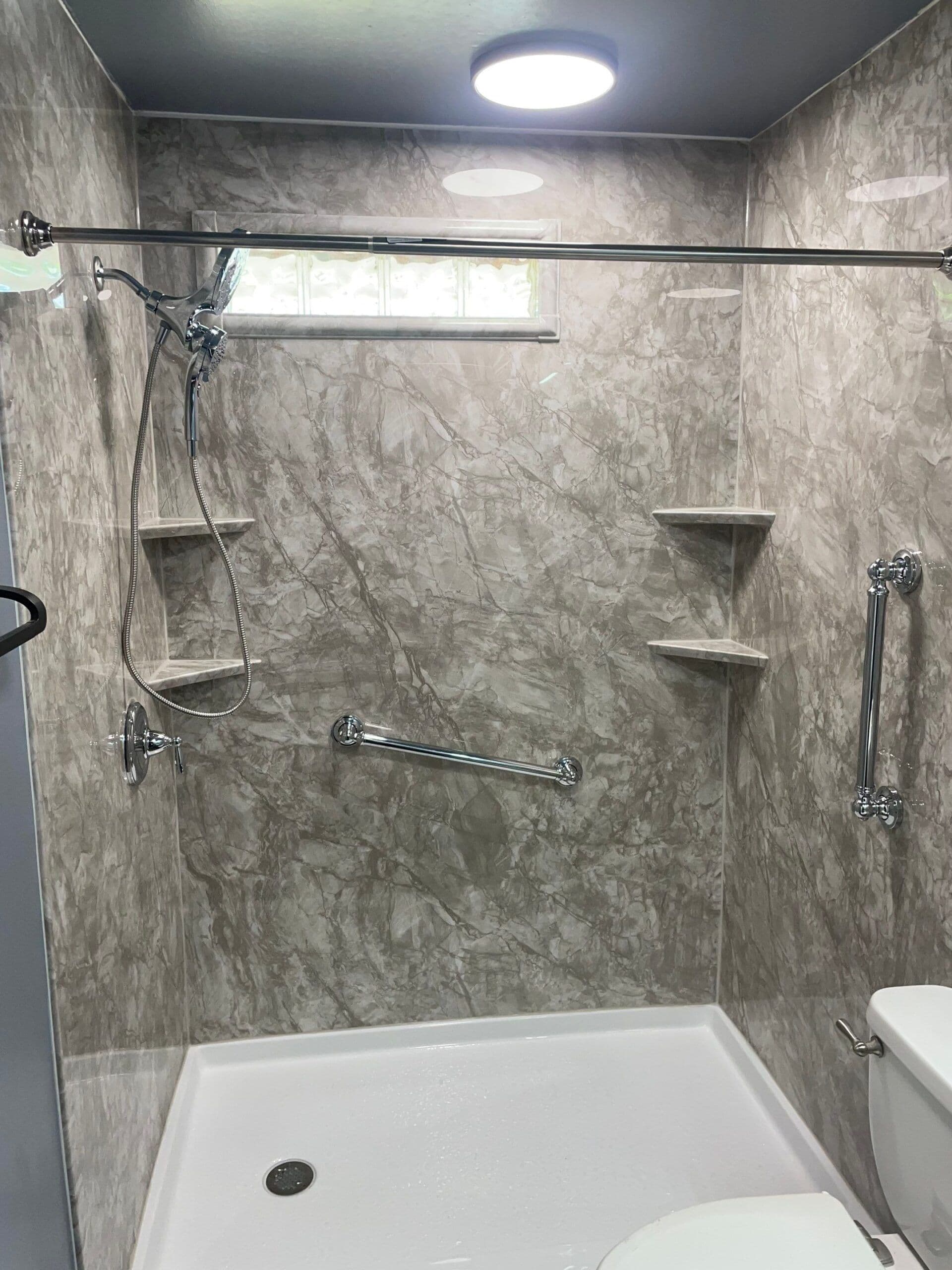 A walk-in shower with gray marbled acrylic walls, multiple grab bars, corner shelves, and small window.