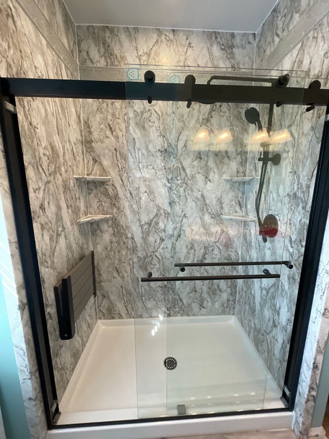 A walk-in shower with sliding glass doors, black fixtures, gray marbled walls, and folding bench seat.