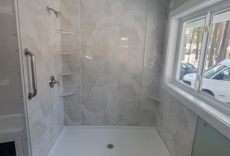 A newly-remodeled walk-in shower with a window to the right, white marbled and textured pattern walls, corner shelves, and vertical safety grab bar.