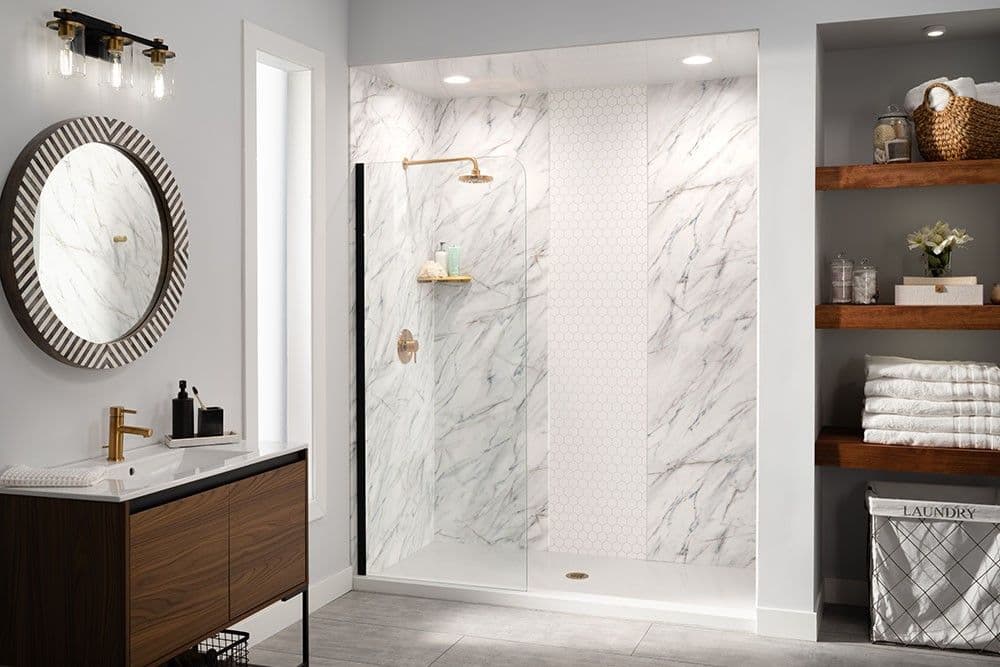 A light gray bathroom with a circular mirror, walk in shower with a glass partition, marbled and hexagonal patterned walls, and gold fixtures.