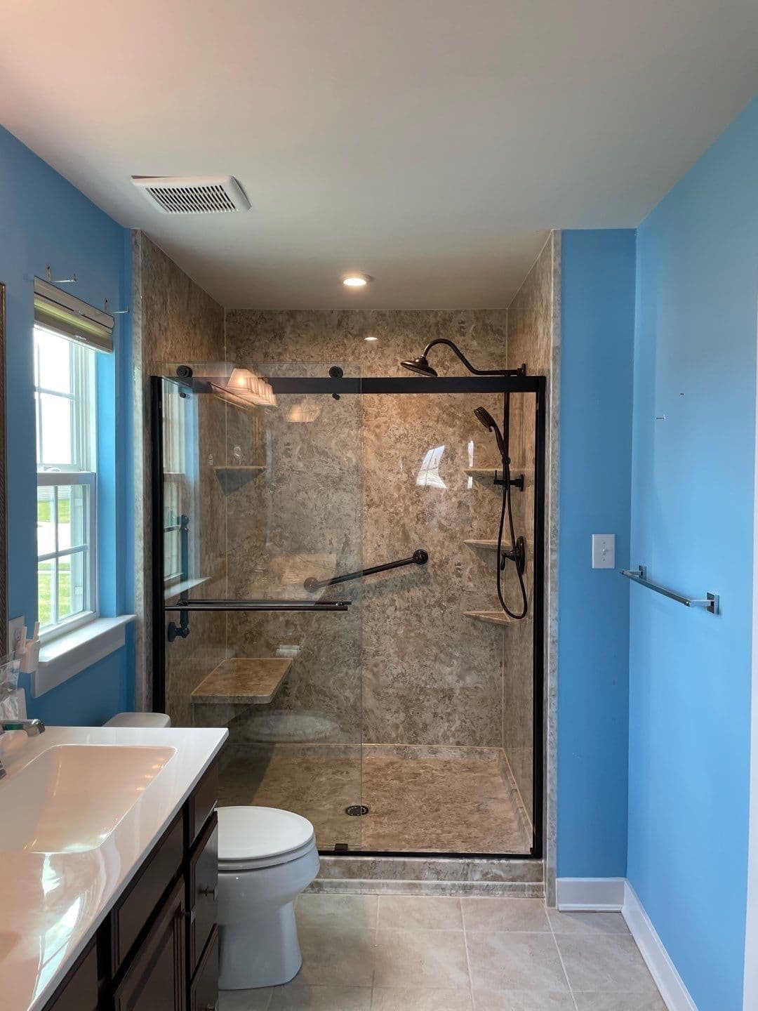 A renovated blue bathroom with a walk-in shower with sliding glass doors, brown marbled walls, black fixtures and grab bar, built-in shower bench and corner shelves.