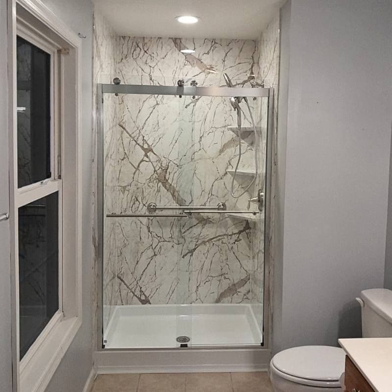 A gray bathroom with a converted tub to walk-in shower with beige and white marbled walls, sliding glass doors, and grab bar.