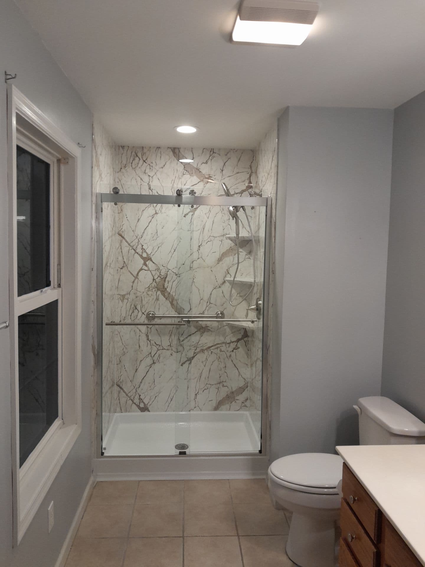 A gray bathroom with a converted tub to walk-in shower with beige and white marbled walls, sliding glass doors, and grab bar.