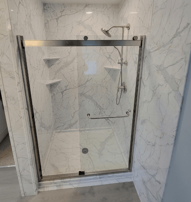 A walk-in shower with sliding glass doors, corner shelves, and marbled walls.