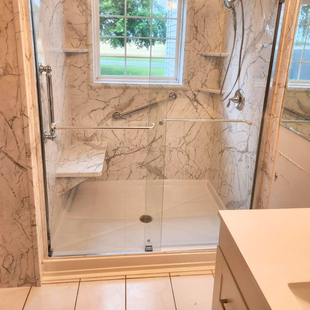 A walk-in shower with a built-in shower bench, white and gray marbled walls, corner shelves, and multiple safety grab bars.