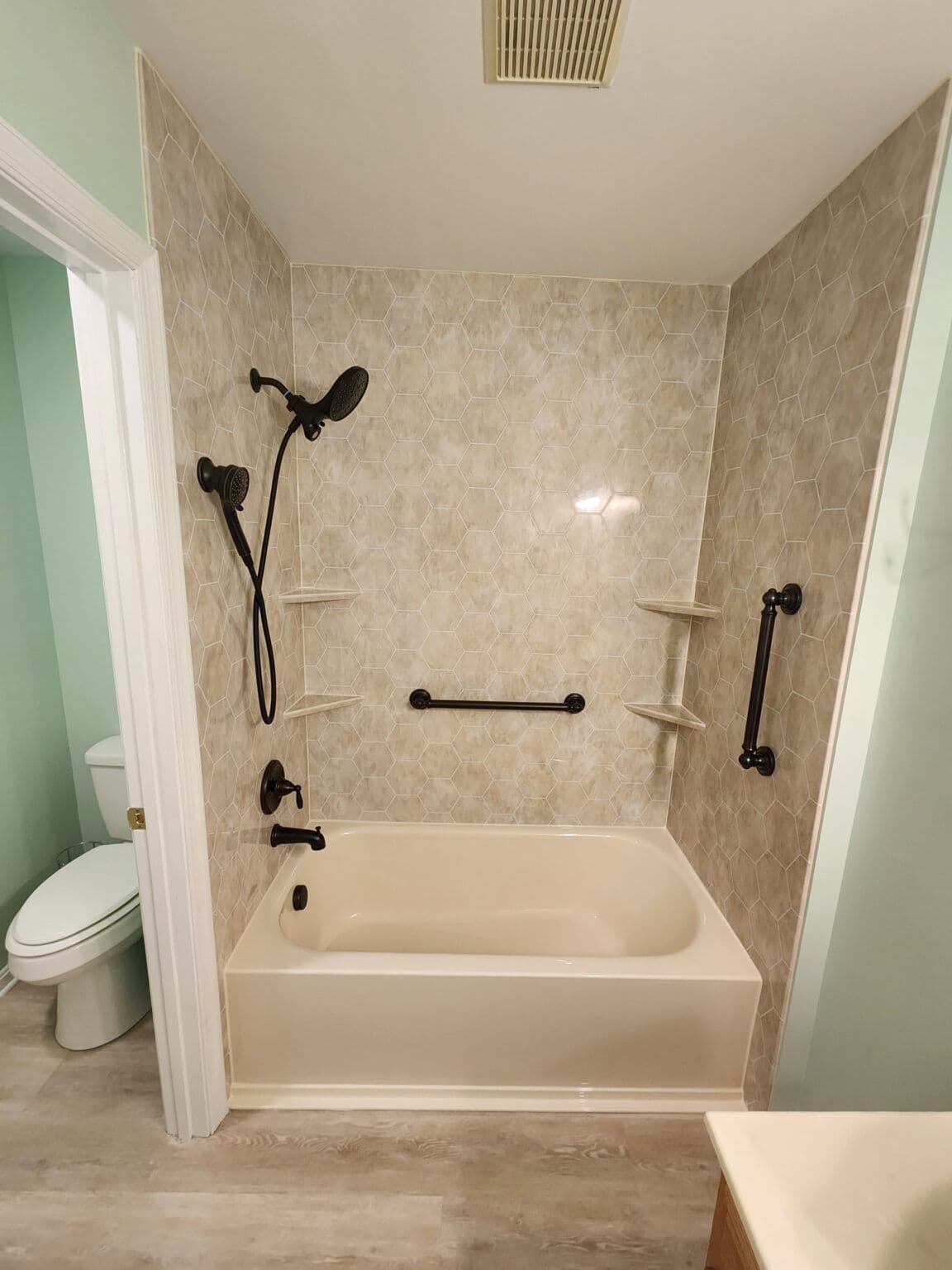 A green bathroom with combination shower tub with black fixtures, two grab bars, brown hexagonal patterned walls, and corner shelves.