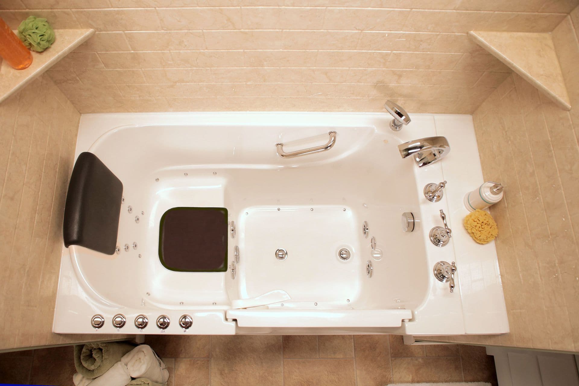 A top-down view of a walk-in bathtub with built-in grab bar, beige tile walls, and corner shelves.