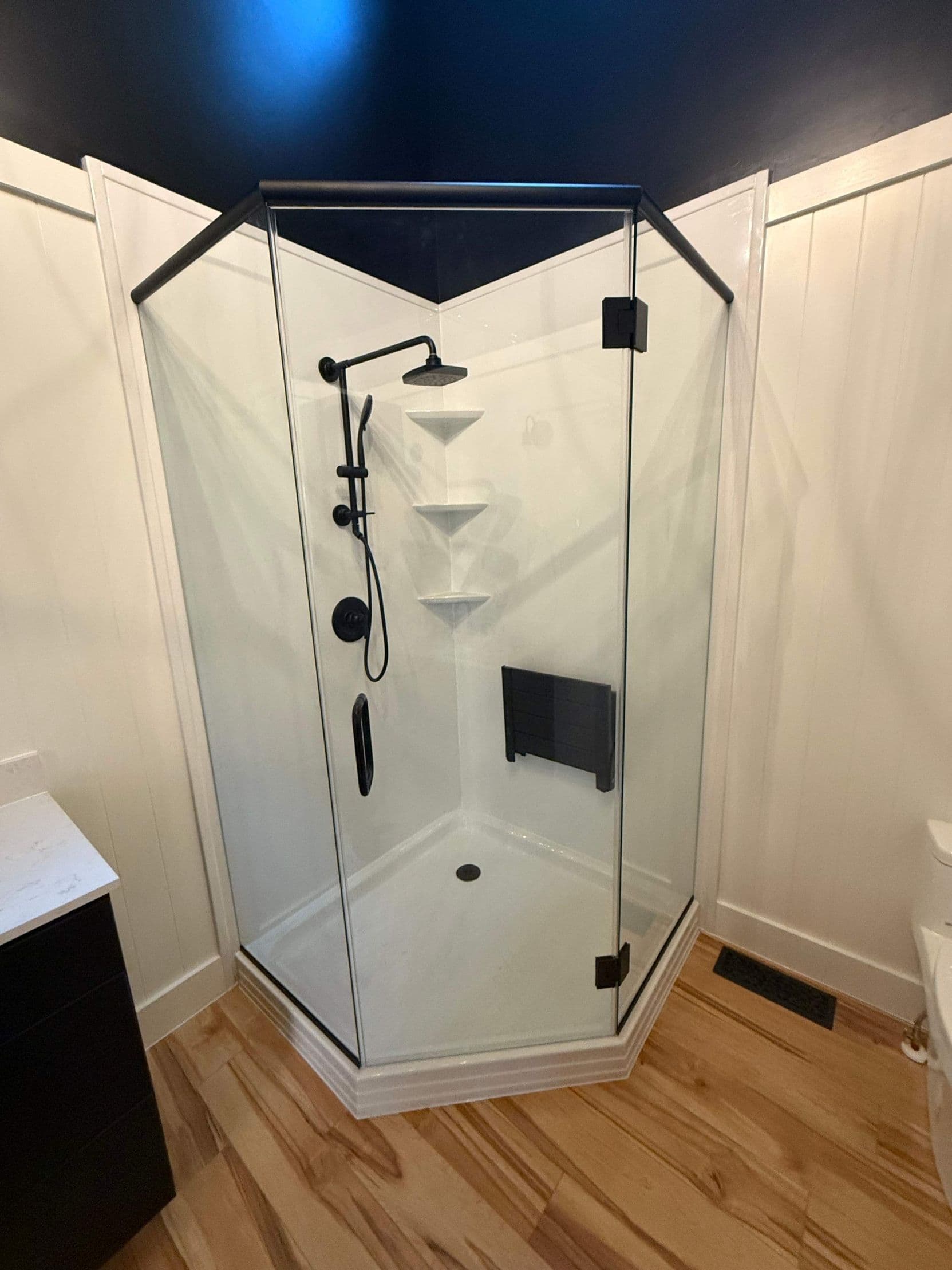A corner shower with glass walls, black fixtures, folding shower bench, and corner shelves.
