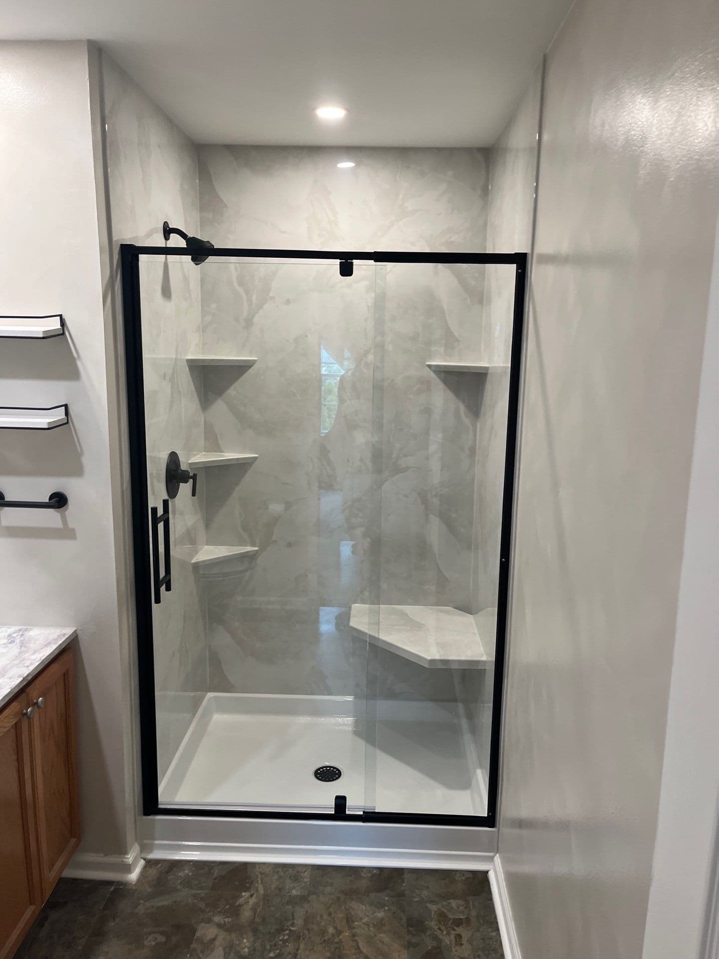 A walk-in shower with swinging glass door, black fixtures, corner shelves, and built-in bench.