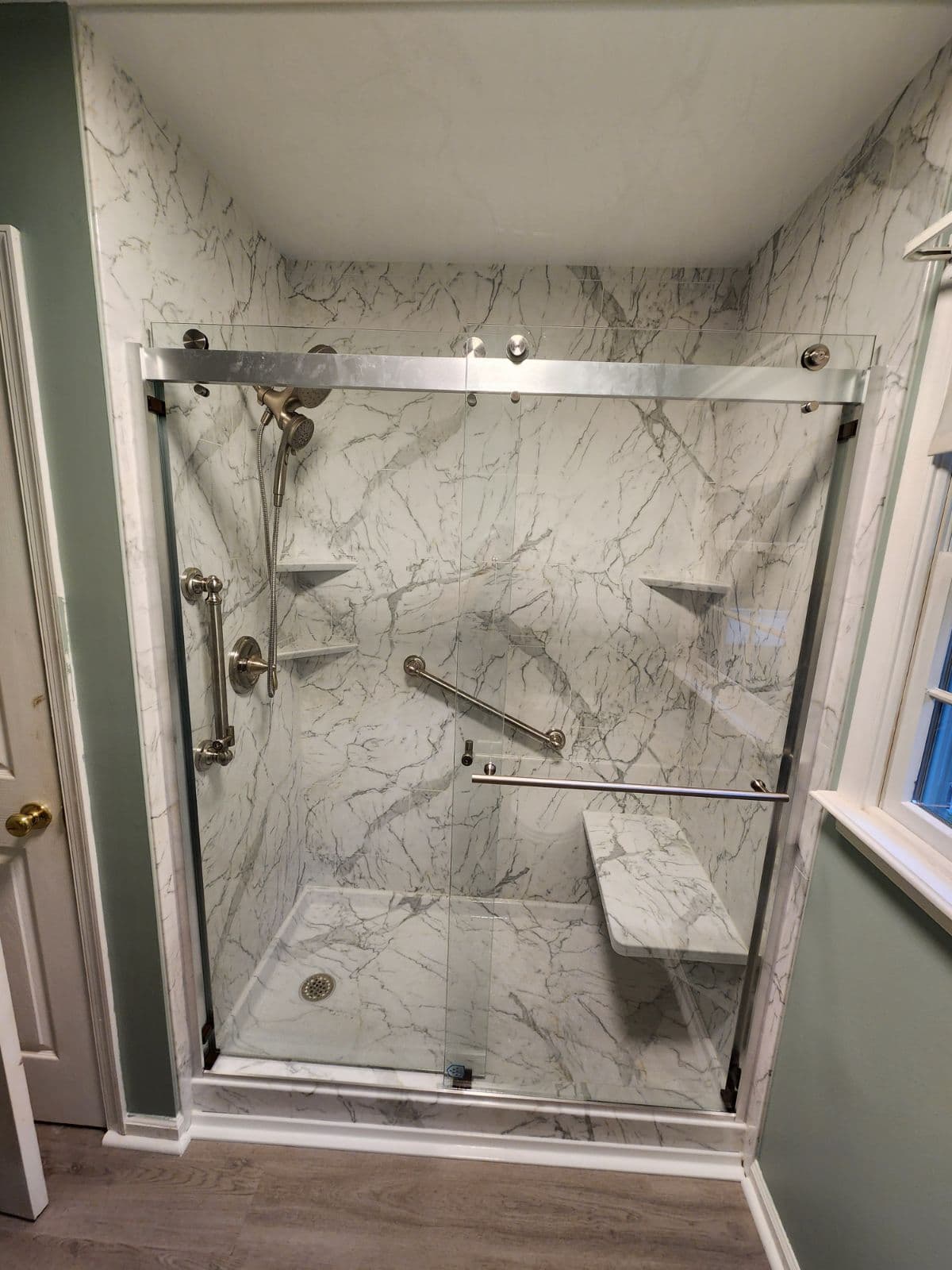 A walk-in shower with sliding glass doors, built-in bench, grab bars, and marbled walls.