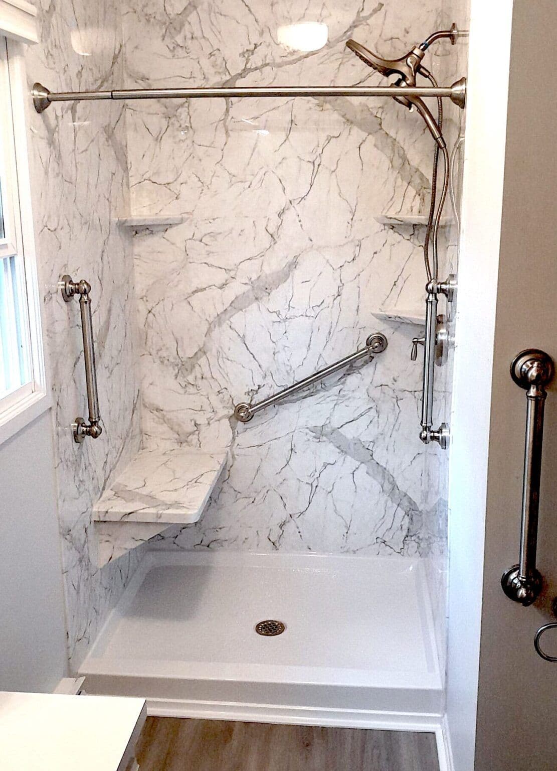 A walk-in shower with a built-in shower bench, white and gray marbled walls, corner shelves, and multiple safety grab bars.