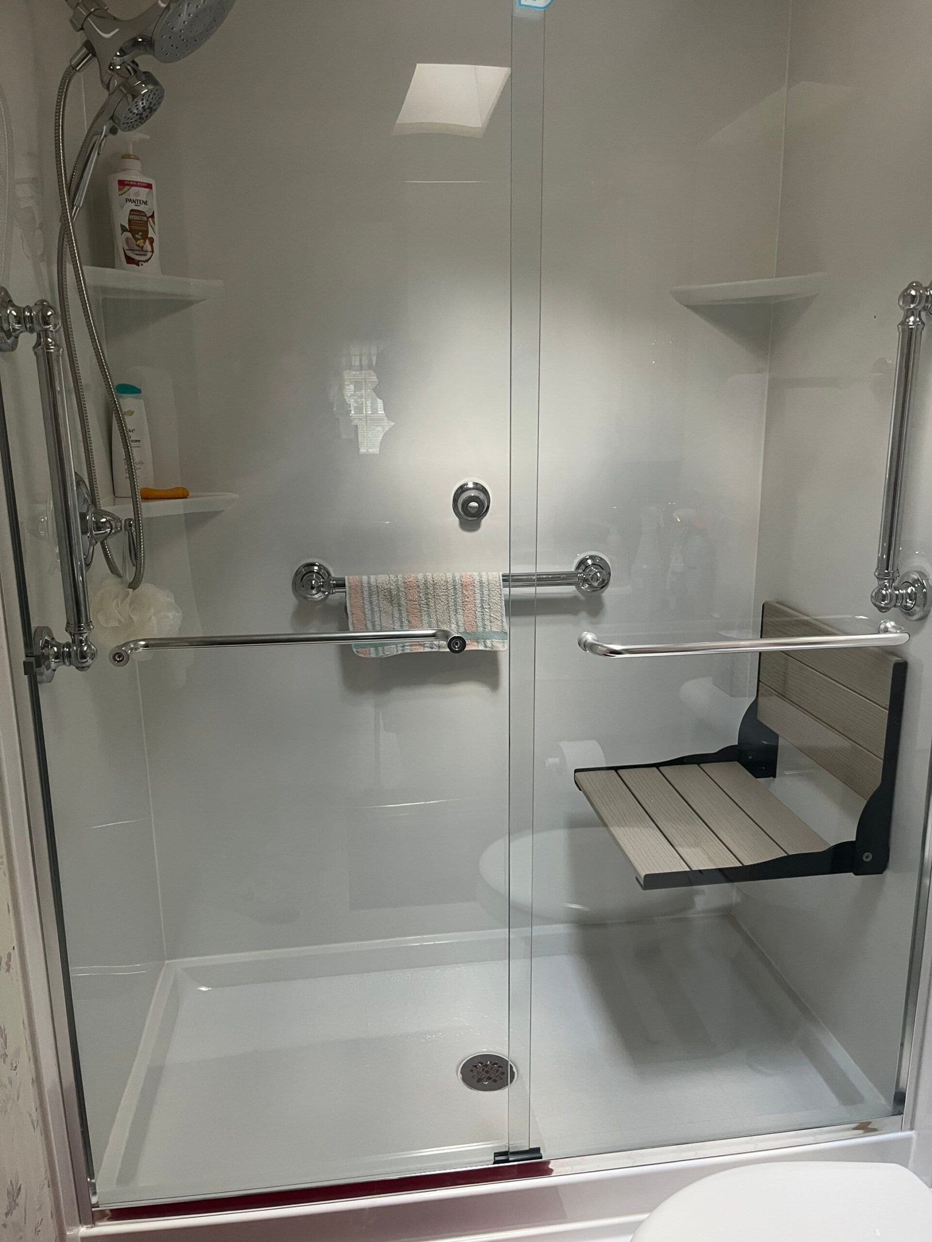 An accessible walk-in shower with sliding glass doors, folding bench seat, grab bars, and corner shelves.