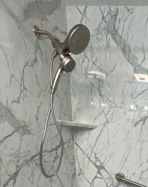 A shower with detachable showerhead in a shower with white and gray marbled walls and corner shelves.