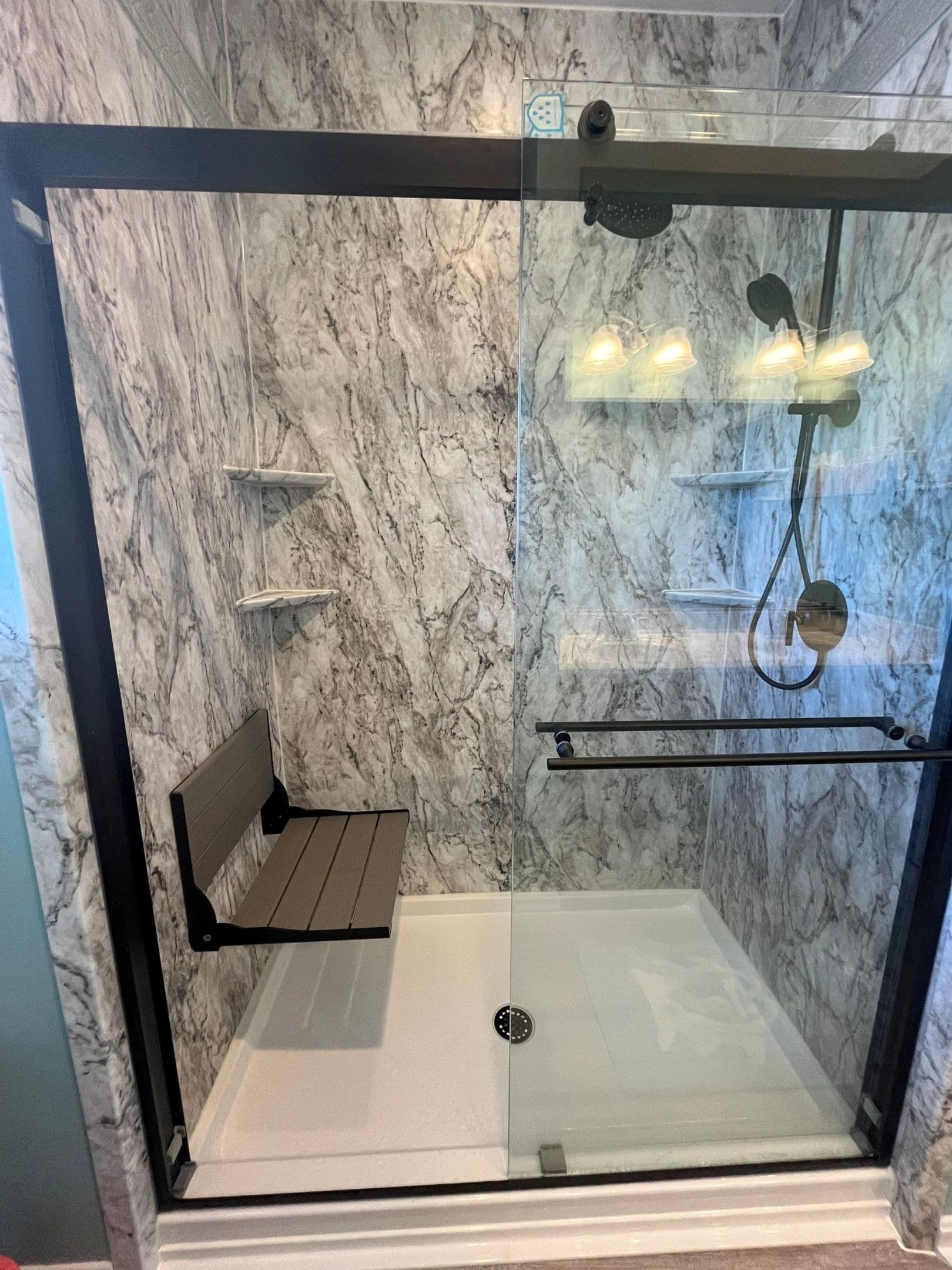 A walk-in shower with sliding glass doors, black fixtures, gray marbled walls, and folding bench seat.
