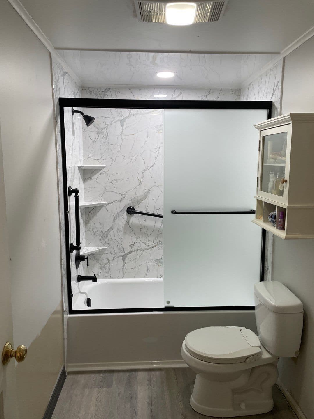A combination shower tub with sliding frosted glass doors, corner shelves, and gray and white marbled walls.