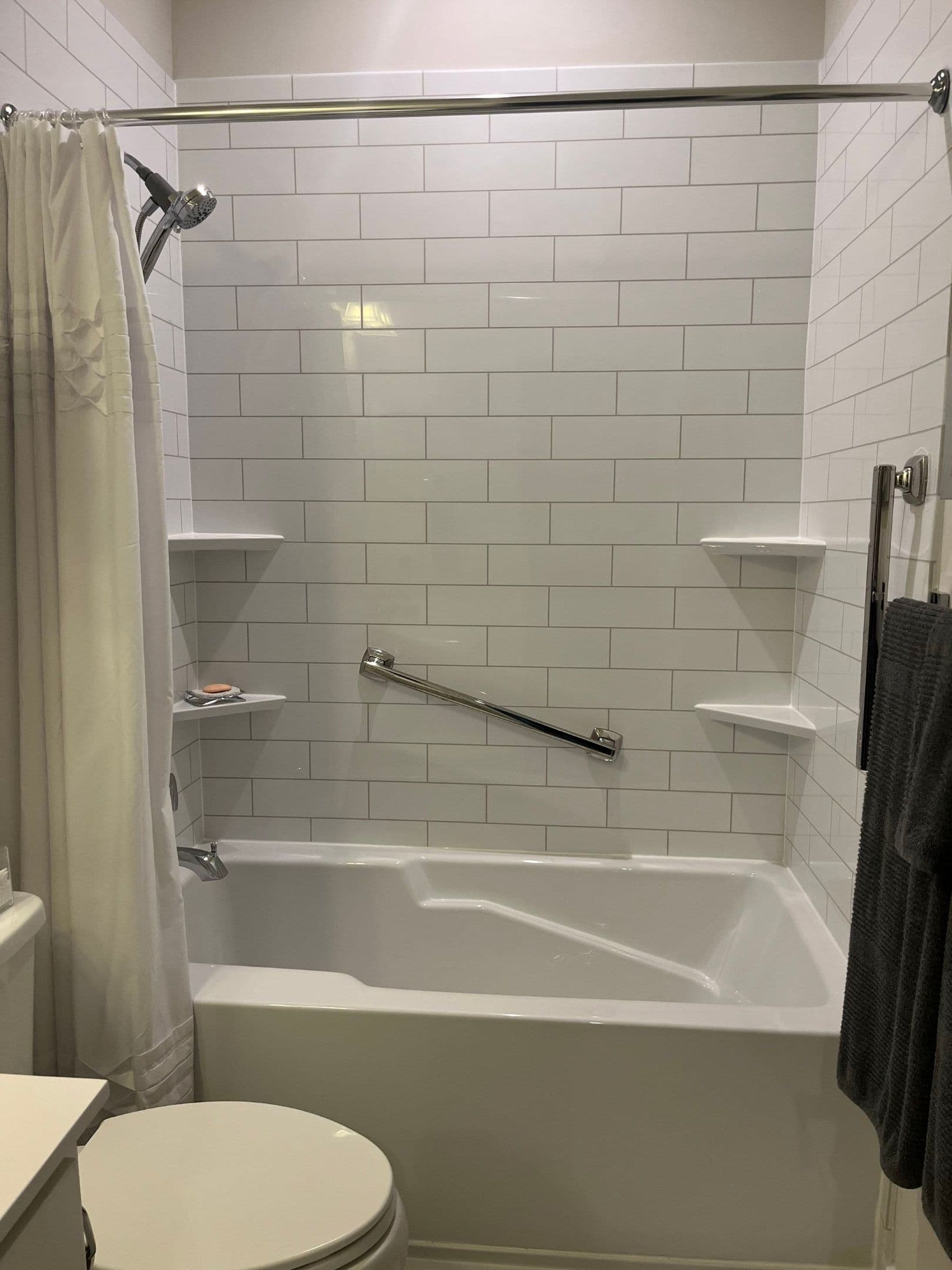 A combination shower tub with white tile pattern walls, corner shelves, and diagonal safety grab bar.