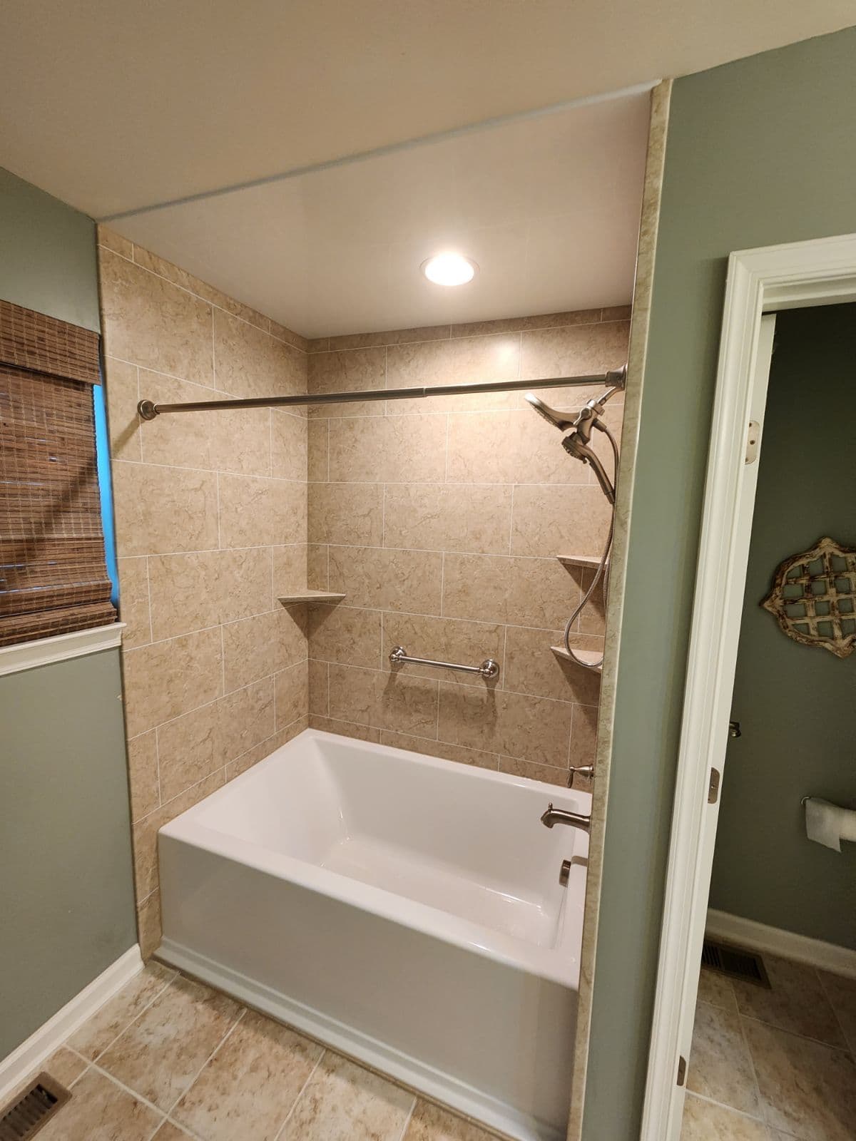 A small green bathroom with a remodeled combination shower tub with beige marbled walls, grab bar, and corner shelves.