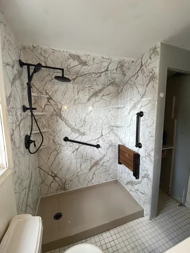 A walk-in shower with tan shower pan, black fixtures, grab bars, built-in bench, rainfall showerhead, and marbled walls.