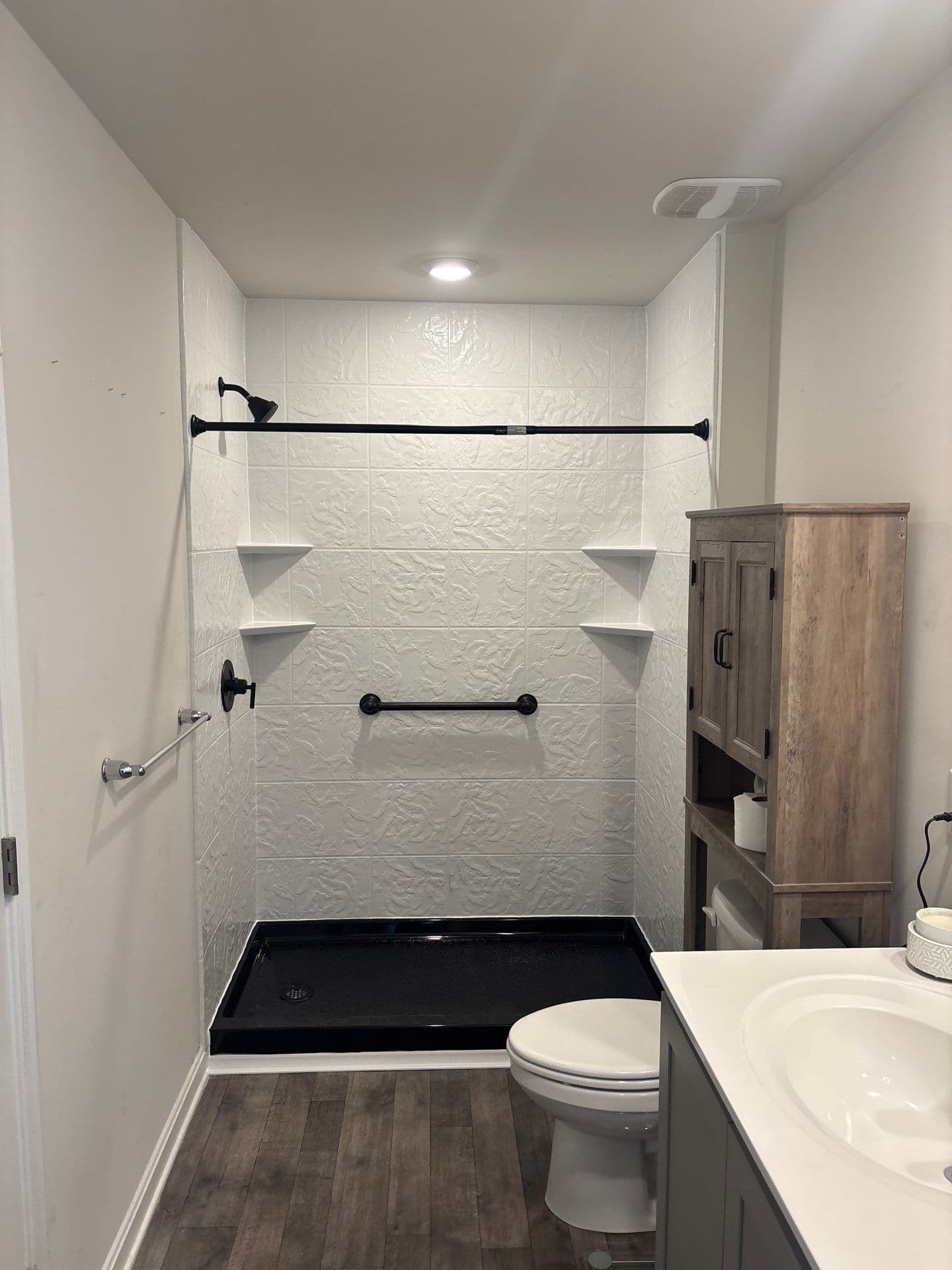 A walk-in shower with black base and fixtures, grab bar, white textured walls and corner shelves.